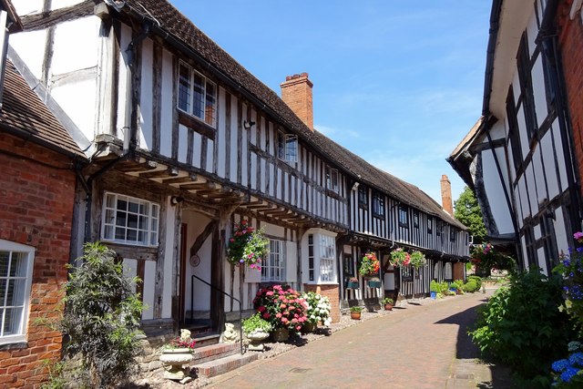 Row of historic half-timbered cottages with white plaster and dark timber framing along a sunny brick-paved street, with hanging flower baskets and potted plants in bloom.