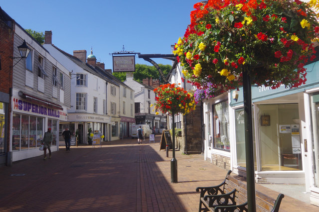 Sunlit pedestrian street in a small town with white storefronts, brick pavement, hanging flower baskets, and people strolling.