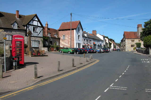 Sunny village street with classic English cottages, parked cars, and a red roadside telephone booth on the left.