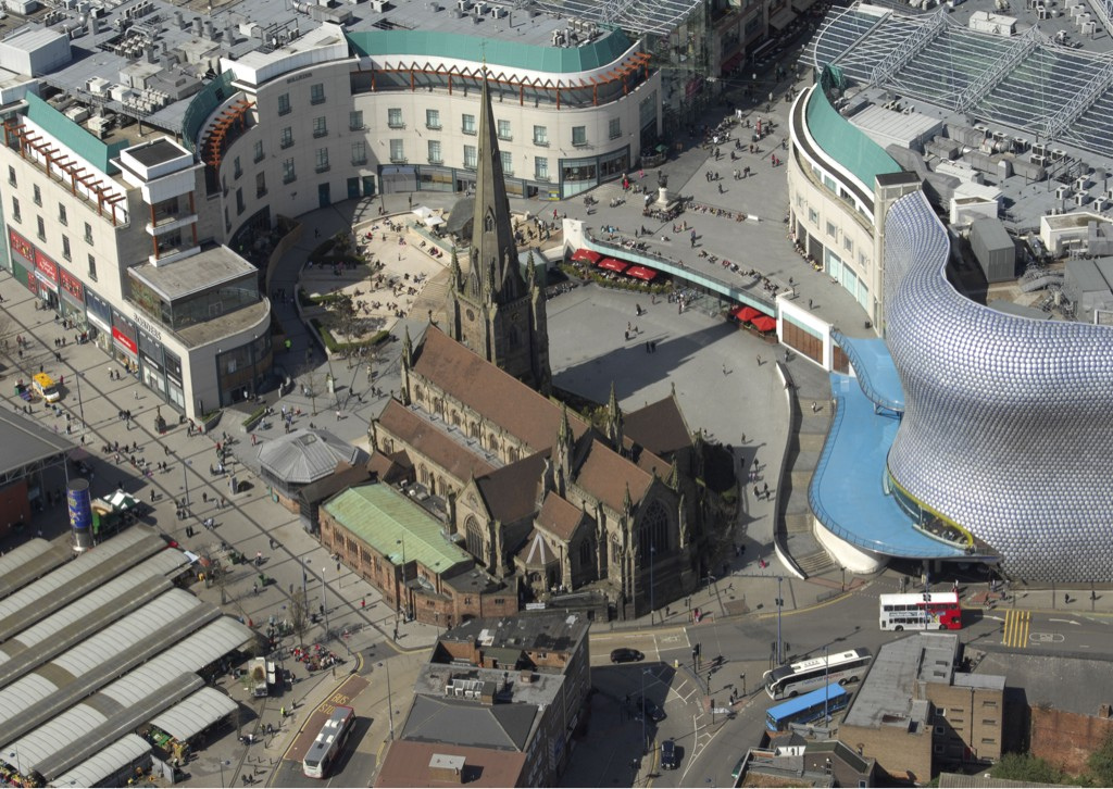 Aerial view of a busy city plaza featuring a medieval church surrounded by modern curved buildings and pedestrian areas.