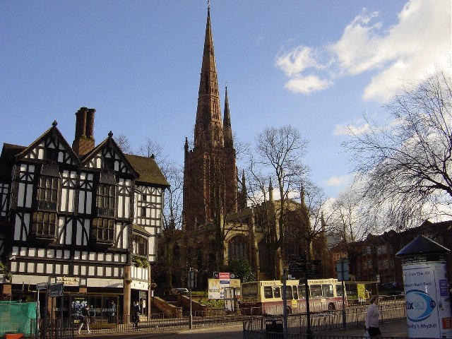 A tall Gothic church spire rises behind a row of black-and-white timber-framed houses along a street, with buses and bare trees in the foreground.