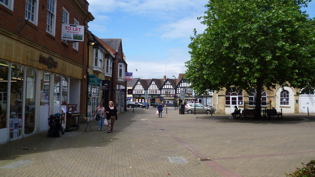 Pedestrian shopping street with brick storefronts, people walking, and a large tree on the right under a blue sky.