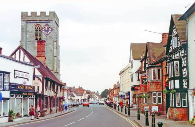 Historic town street with a tall stone church tower on the left, timber-framed shopfronts, and red-brick buildings lining a curved road with pedestrians and cars.