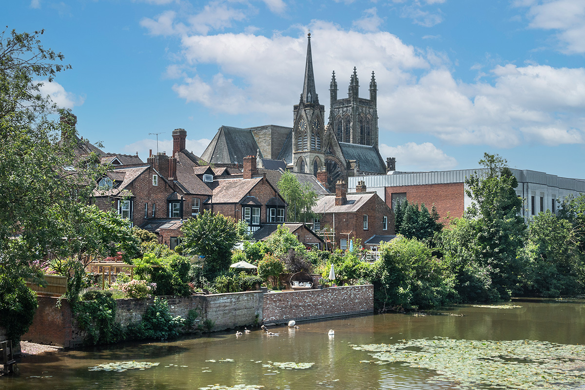 Riverside town with brick houses, a tall Gothic church, and green gardens along the water under a blue sky.