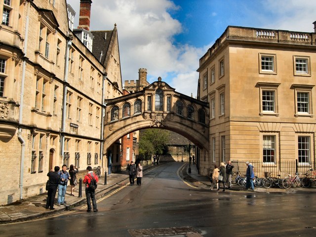 Stone historic arch bridge linking two beige stone buildings over a street, pedestrians walking and cyclists parked nearby on a sunny day