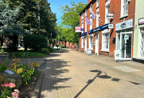 Sunny pedestrian street lined with brick storefronts and banners, trees on the left and a lamppost shadow on the pavement.