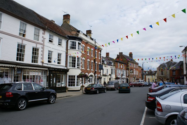 Quaint town street with colorful pennant flags overhead, row of brick and plaster storefronts, and parked cars along the curb.