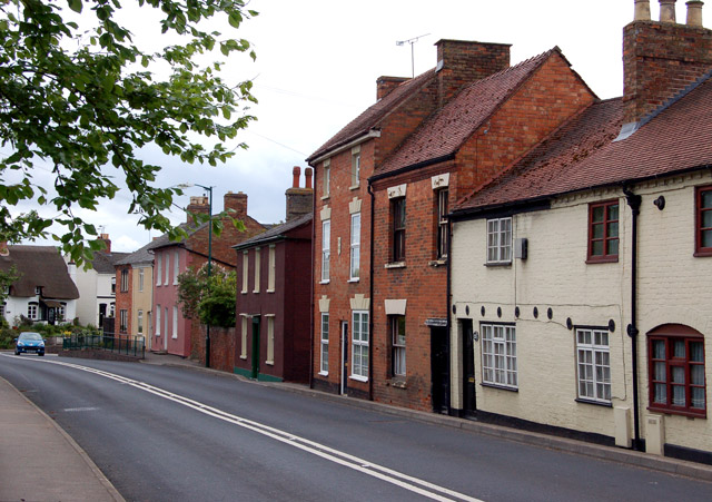 Row of colorful brick terraced houses along a quiet residential street with a tree overhead and double white road lines in the foreground.