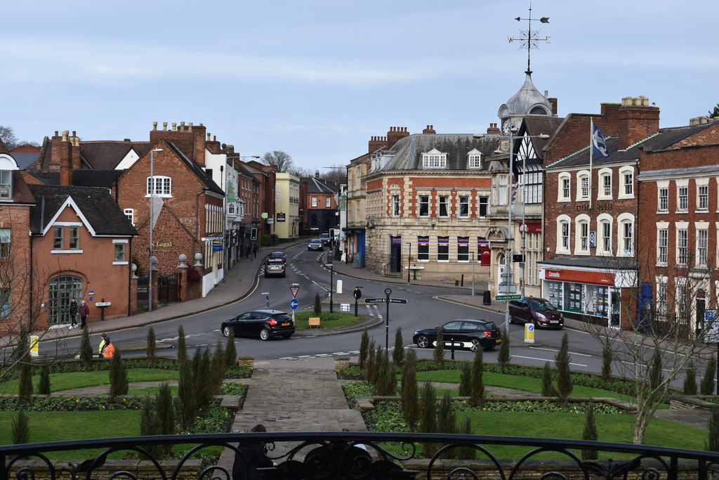 View of a small town street with brick buildings, a roundabout, and a stone path from a central garden foreground.