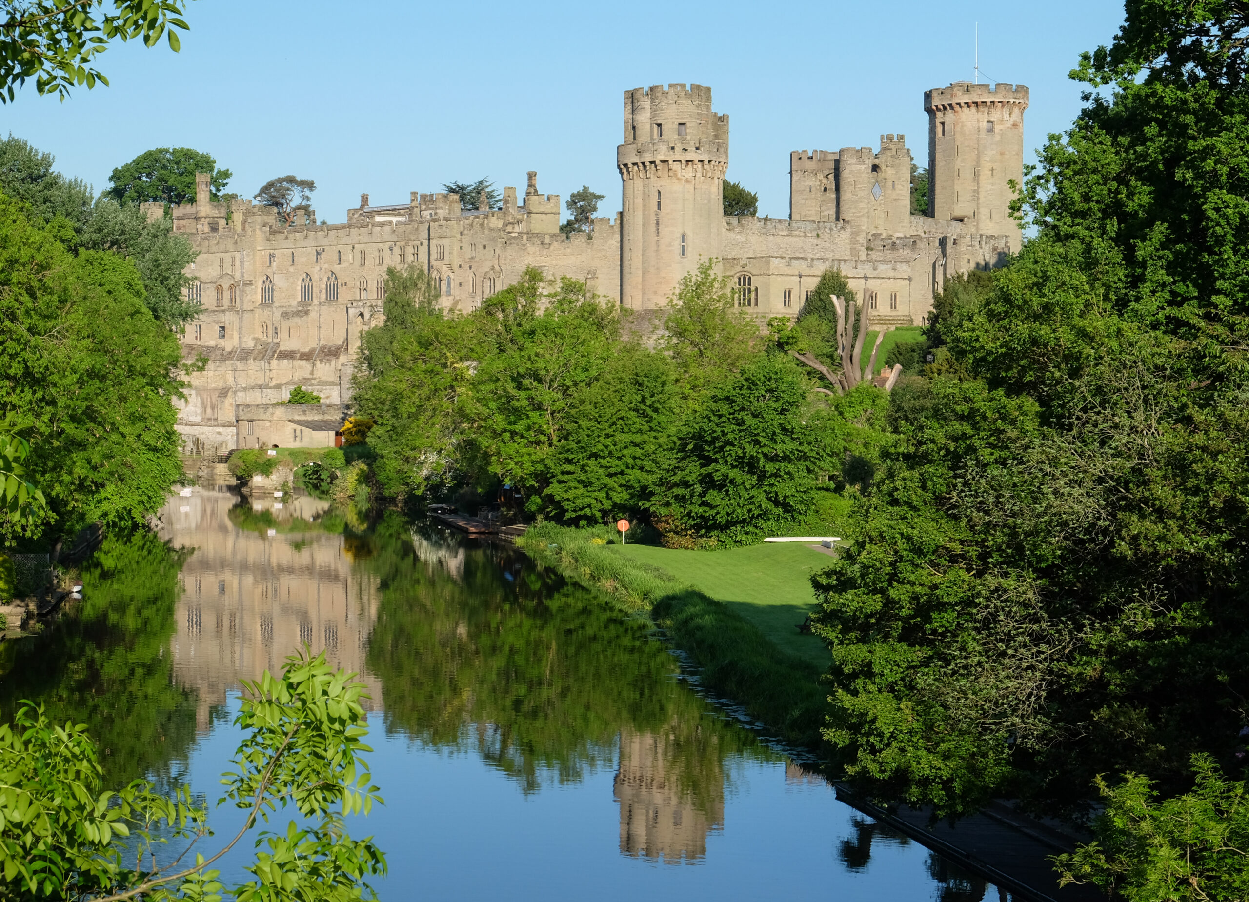Castle with tall rounded towers along a calm river, surrounded by green trees on a sunny day.