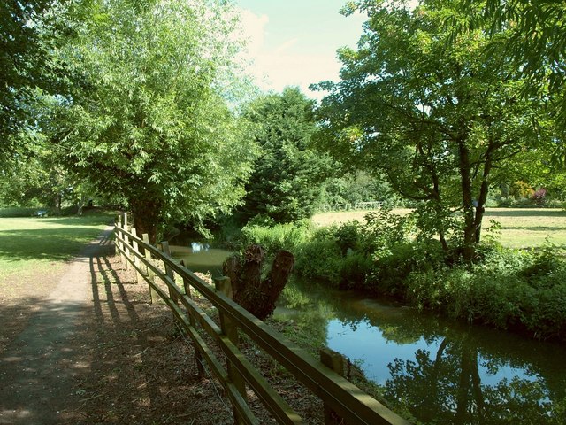 Sunlit park path beside a calm canal with a wooden fence and green trees in the background.
