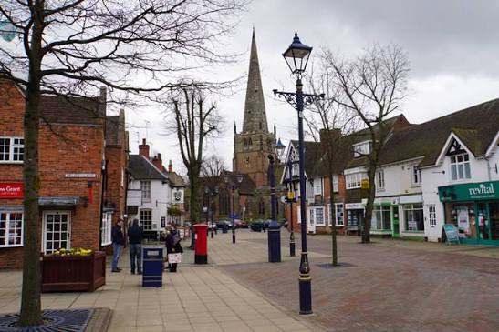 Town street with a distant church spire, leafless trees, pedestrians, a red postbox, and shops lining both sides.